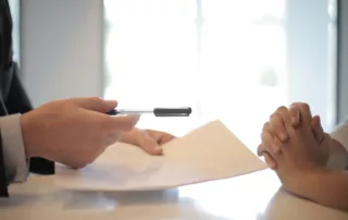 A white business man holding a pen and paper, handing it to a business woman to sign.