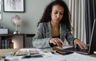 A black business woman reviews her cash and accounting with a laptop and calculator.