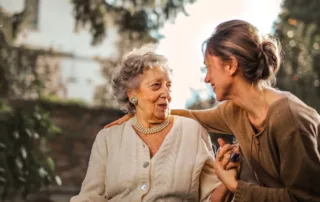 A caregiver holds an elderly woman’s hand while she smiles.