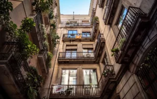 Looking up into a tall, older style apartment building with balconies full of potted plants.