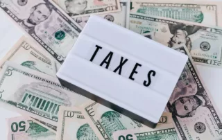 A white sign that reads “taxes” laying on top of dollar bills.