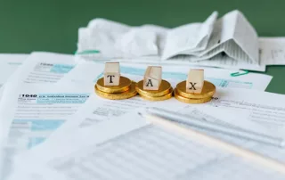 Tax Documents on the Table with gold coins stacked on top and wooden blocks spelling out "TAX".