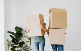 Two tenants holding moving boxes over their faces inside a new, empty apartment.