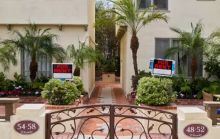 Apartment building entrance with For Rent signs on the front lawn, featuring a charming brick pathway, tropical palm trees, and an elegant wrought iron gate, real estate rental concept.